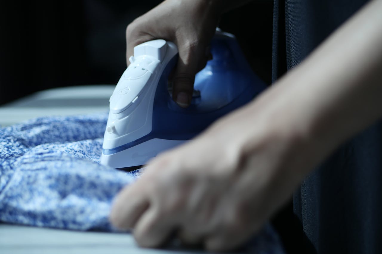 Detailed view of hands using an electric iron on blue fabric indoors.