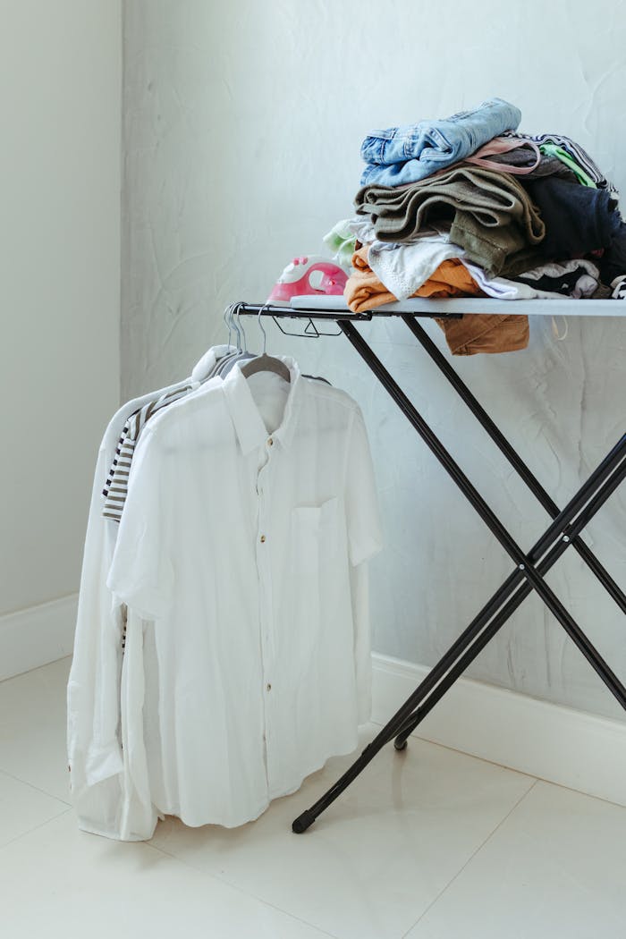 Neatly folded clothes on ironing board with hanging shirts in a tidy laundry setup.