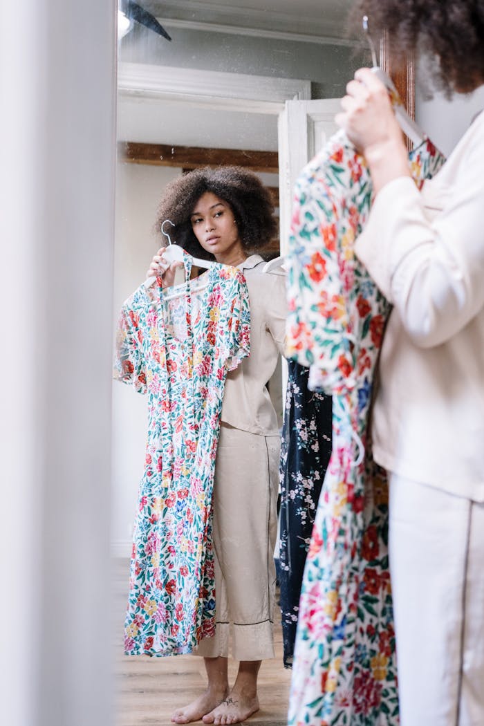 A woman with curly hair choosing floral dresses in front of a mirror at home during morning routine.